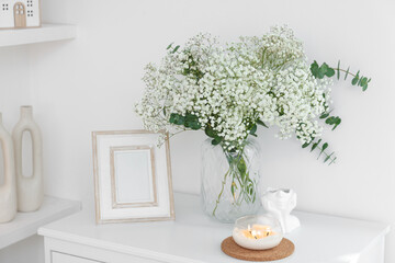 Bouquet of beautiful gypsophila flowers and eucalyptus branches in vase surrounded by decorative elements on chest of drawers indoors