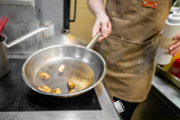 A chef in an apron cooks chopped vegetables in a pan on a stove. Steam rises, emphasizing the culinary process in a professional kitchen setting.