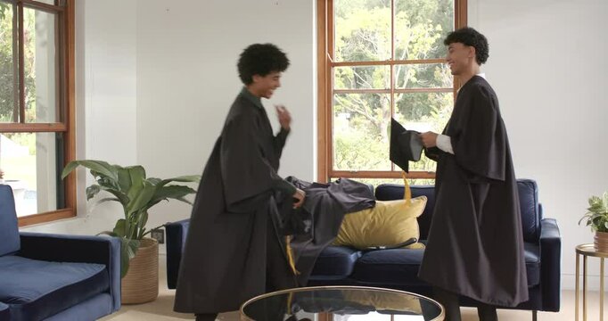 Proud father congratulating twin diverse sons in graduation gowns at home celebration