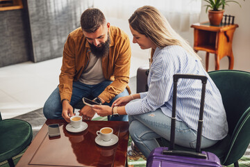 Cheerful adult couple is going on vacation. Tourists sitting in hotel lobby, drinking coffee and using smartphone.