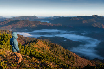 Fototapeta premium Rear view of a young man wearing a blue jacket, gazing at the breathtaking view from the top of a mountain at sunrise, with low clouds filling the valley below
