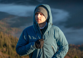 Portrait of a serious man with stubble, wearing a blue hooded jacket, standing in the mountains,...