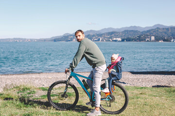 Fototapeta premium A father riding a bicycle with his young child seated in a safety seat on the back. They are enjoying a scenic outdoor ride near a beautiful lake with mountains in the background on a sunny day