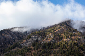 Fototapeta premium Scenic mountain view with misty clouds rolling over evergreen forests and rugged terrain, creating a dramatic and peaceful atmosphere.