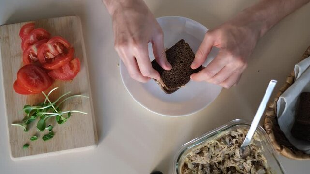 A young  man is preparing a sandwich with fish pate, herbs and tomatoes on rye bread