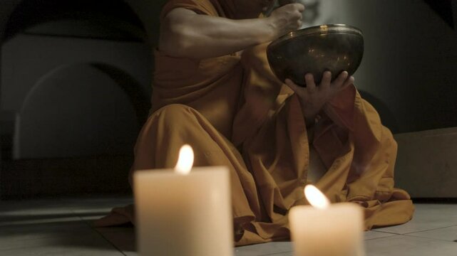 A monk in an orange cassock sits on the floor and runs a hammer along the edges of a beautiful metal singing bowl against a background of burning candles. Indoor sound therapy.