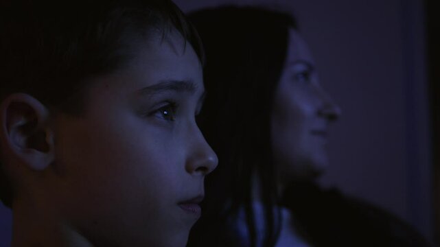 Mother and child watching TV together cozy dark room. Close-up faces lit by blue screen light. Family time, bonding moment, shared entertainment. Parent and kid, home evening, television viewing.