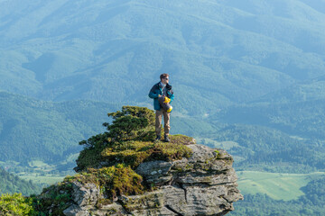 A man hiker puts clothes into a duffel bag while standing on the edge of a rocky mountain peak at sunset, with a breathtaking view of the landscape.