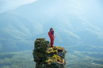 Obraz premium Young European man standing in a red sleeping bag on the edge of a rocky mountain peak, surrounded by mountain ridges.