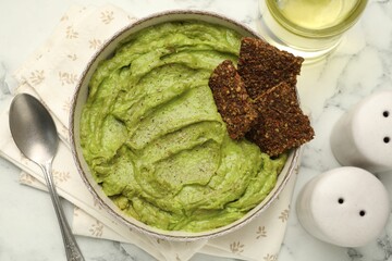 Tasty avocado dip in bowl, crispbreads and oil on white marble table, flat lay