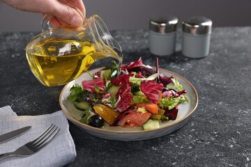 Woman pouring oil onto salad at dark textured table against grey background, closeup