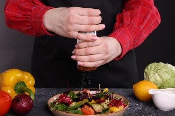Woman salting tasty salad at dark textured table, closeup
