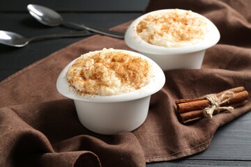 Delicious rice pudding with cinnamon in bowls and spoons on black wooden table, closeup