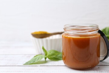 Tasty curry sauce in glass jar, powder and basil on white wooden table, closeup