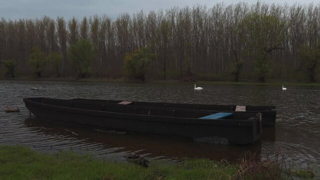 Wooden Boat Docked by River Bank in Wintertime with Bare Trees and Swan in Background

