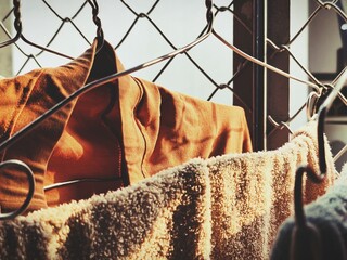 A warm-toned image of an orange shirt and a beige towel hanging on hooks against a wire fence, lit by soft sunlight.