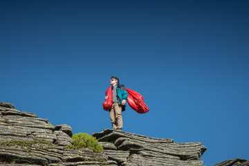 Young man wearing sunglasses and outdoor gear standing on a rocky mountain peak, holding a red...