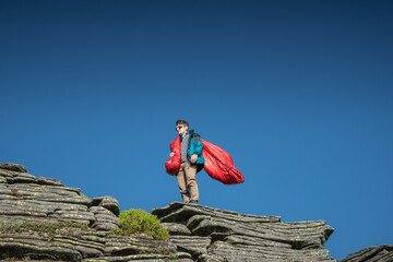 Young man wearing sunglasses and outdoor gear standing on a rocky mountain peak, holding a red...
