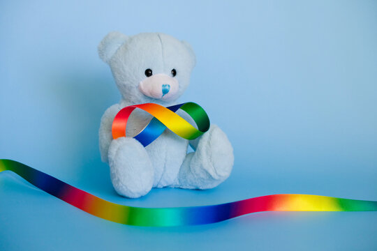Autism acceptance month, Autism awareness day concept. Toy teddy bear with rainbow satin ribbons in shape of infinity on blue background. Infinity is symbol of autism disorder, ADHD, neurodiversity