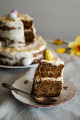 Piece of homemade two layered Easter carrot cake with ricotta cream, decorated with chocolate eggs and almonds on ornate plate and daffodils on linen tablecloth.