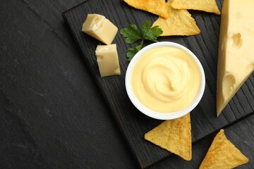 Tasty dipping cheese sauce in bowl, nacho chips and parsley on black table, top view. Space for text