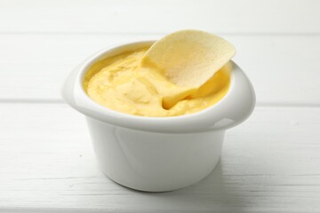 Tasty dipping cheese sauce in bowl and potato chip on white wooden table, closeup