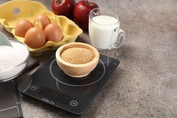 Digital kitchen scale with bowl of brown sugar and other products on grey table, closeup. Space for text