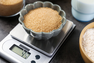 Digital kitchen scale with bowl of brown sugar and other products on grey table, closeup