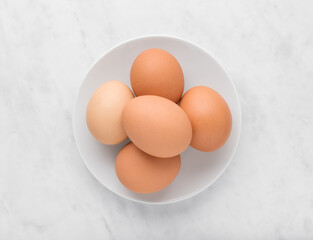 Chicken eggs in a white ceramic bowl on marble background, top view