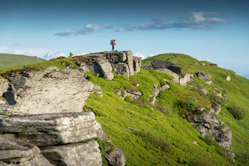 Female hiker wearing a red jacket and an orange beanie, with trekking poles and a backpack, hiking on a rocky mountain summit.