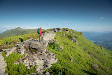 Female hiker wearing a red jacket and an orange beanie, with trekking poles and a backpack, stands on a rocky mountain summit, gazing into the distance