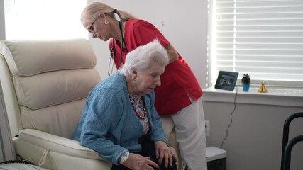 A mature woman in her 80s or 90s is receiving support from a caregiver in a well-lit living room. The caregiver is gently assisting her while standing beside a mobility aid.