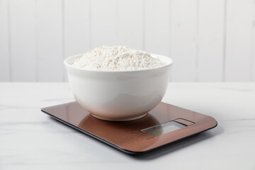 Modern electronic kitchen scale with bowl of flour on white marble table, closeup