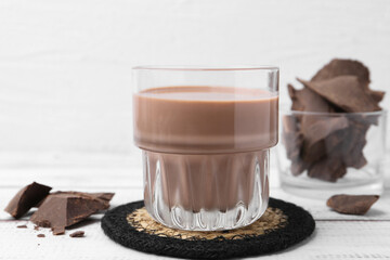 Tasty chocolate milk in glass and pieces of chocolate on white wooden table, closeup