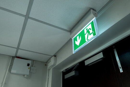 Industry-standard illuminated Fire Exit sign seen at the fire escape and emergency exit doors within a typical hospital. An alarm system can be seen in the background.
