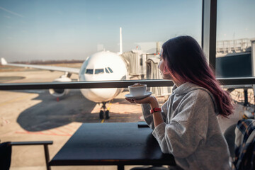 Passenger drinking coffee looking at airplane through window at airport cafe while waiting for...