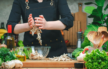 Chef pours chopped oyster mushrooms in bowl.  Cozy kitchen with wooden table, kitchenware, vegetables, herbs and ingredients for cooking. Healthy vegan food, culinary, recipes, blogging