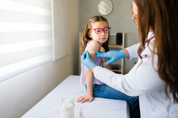 Pediatrician applying bandage to girl's arm after administering a vaccine in a medical clinic,...