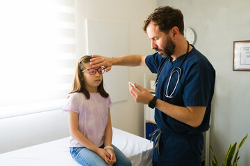 Male pediatrician using digital thermometer checking forehead temperature of ill girl patient...