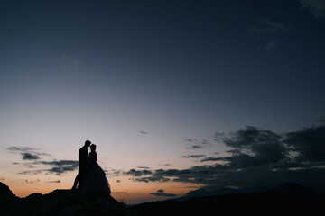 Silhouettes of the bride and groom standing on the mountain and almost kissing at dusk