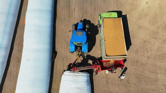Aerial overhead shot on Plastic bag silos and wheat grinding, Milling. Tractor with trailer working in tandem alongside a working combine harvester discharging grain from uploader in an cornfield