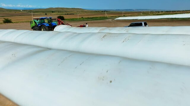 Aerial overhead shot on Plastic bag silos and wheat grinding, Milling. Tractor with trailer working in tandem alongside a working combine harvester discharging grain from uploader in an cornfield