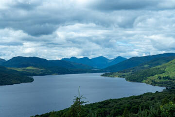 A serene Loch Katrine in Scotland, surrounded by green forests and rolling hills under a dramatic cloudy sky, showcasing the natural beauty of the Highlands and a peaceful pristine landscape - UK