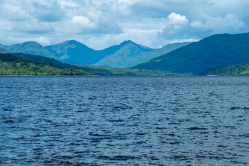 Picturesque view of Loch Katrine surrounded by lush green mountains under a partly cloudy sky, offering a serene and natural landscape ideal for exploration and outdoor enthusiasts - Scotland, UK