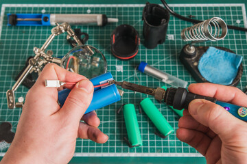 Close-up scene of repairing an old power tool battery on a soldering table with soldering iron, wires, battery cells, and green grid mat — perfect for electronics and DIY content.