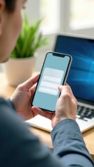 Person using smartphone to interact with mobile application while sitting at desk with laptop and plant in background, digital communication concept