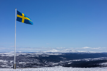 Swedish flag flying high over snowy mountains under a clear blue sky &ndash; a bold symbol of freedom, nature, and northern pride.