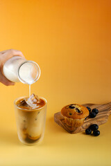 A person is pouring milk into a cup of coffee. The coffee is served on a yellow table with a muffin...