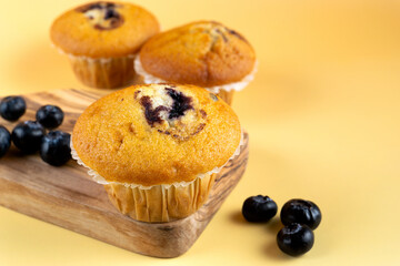 A wooden cutting board with three blueberry muffins and blueberries on it.