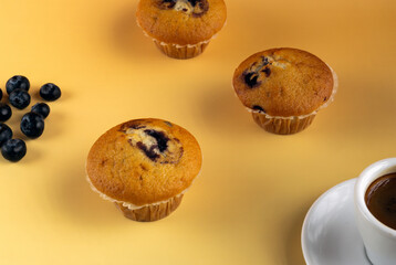 Three muffins with blueberries on a yellow table. A cup of coffee is next to them